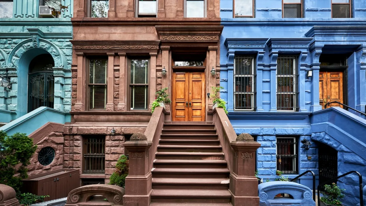 Colorful teal, brown, and blue brownstones in New York City with wide stone stairways going up to first floor doors.